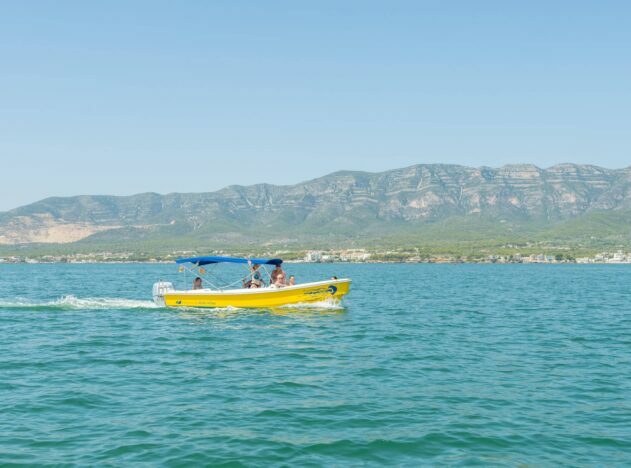 Una llanxa groga amb diversos passatgers, navegant per aigües tranquil·les davant d'una línia de costa amb muntanyes verdes al fons.